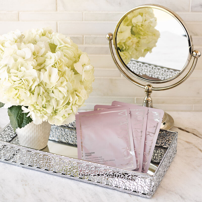 Three packets of TimeWise Repair Lifting Bio-Cellulose Masks from Mary Kay styled in decorative tray with mirror and flowers.