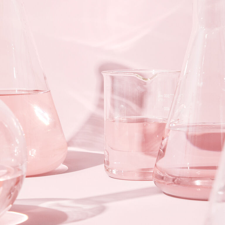 Clear glass beakers with colorless liquid on a pink surface, reflecting soft pink lighting. 