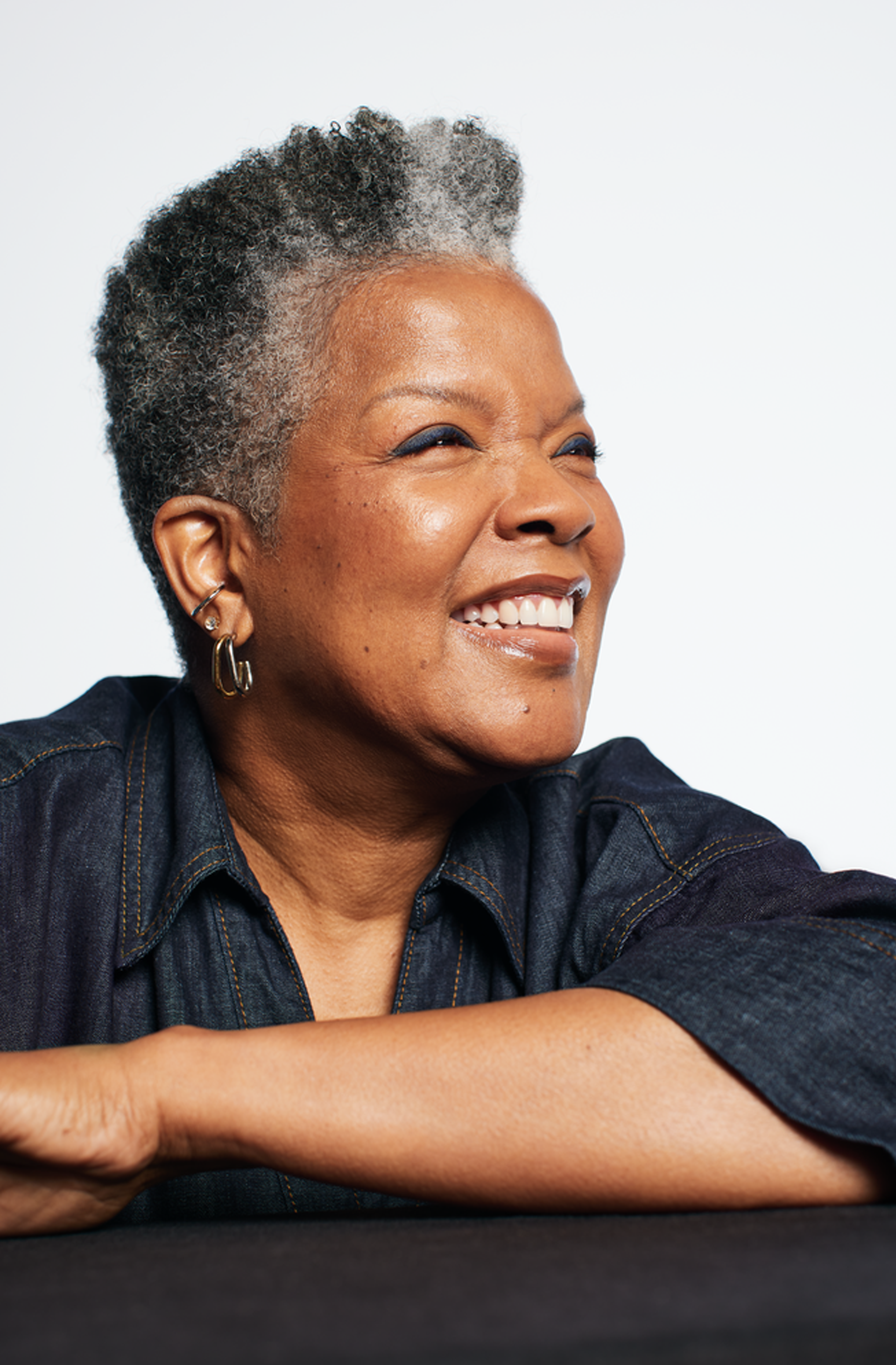 A person with short, textured hair sits with folded arms on a dark surface, wearing a denim shirt and hoop earrings.